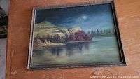 Framed oil painting showing serene lake with mountains, moonlight, trees, teepees, and reflections on water.