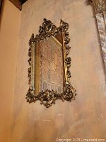 Rococo style rectangular wall mirror with intricate gold leaf frame, detailed scrollwork and floral elements, hung on wall reflecting patterned surface.