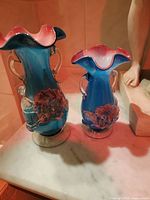 Photo showing two Venetian glass vases with ruffled edges and floral motifs in blue and pink gradient finish on a marble surface, next to a sculpture base and sculpted leg.