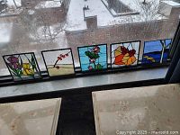 Five small stained glass panels set on a windowsill showing seasonal designs.