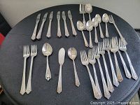 Full set of silver-plated cutlery laid out on black table showing knives, forks, spoons, and spreaders with detailed metal handle designs.