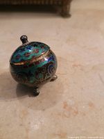 Photo of small round Venetian pill box showcasing cloisonné enamel decoration, hinged lid closed, resting on three metal feet, showing detailed colorful patterns.