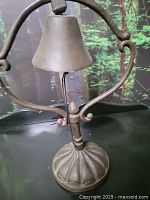 A vintage cast iron farm bell with a leather pull string against a backdrop. Shows the bell suspended on an ornate stand with a fluted base.