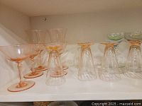 Photo showing pink depression glass stemware, including different styles of glasses such as coupe/wide wine glasses, stem flutes, and champagne glasses on a white shelf.