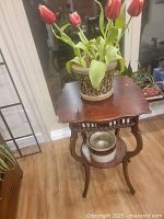 The photo shows a vintage wooden plant table with curved legs and decorative carvings. On top sits a glazed ceramic planter with artificial red tulips. A second ceramic container is placed on the lower shelf of the table.