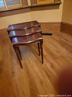 Photo showing a set of three medium-dark varnished wooden nesting tables nested together. Tables have scalloped edges and turned legs with small round feet.