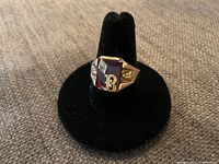 Front view of ring showing red center stone, diamond-cut accent, and gold “B” detail