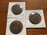 Three one penny bank tokens in protective holders placed on a wooden surface. Visible are the Bank of Upper Canada tokens from 1850 and 1857, and the Province of Canada Bank of Montreal token from 1842.