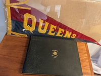 Photo shows black hardcover yearbook with gold Queen's University crest and the year 1924, alongside the vintage Queens school banner partially visible behind it.