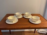 Four shell-shaped tea cups with matching saucers on a wooden table, displayed in square formation.