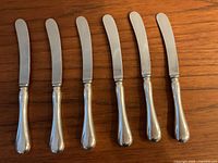Top view of six sterling silver handled butter knives with stainless steel blades arranged in a row showing blade and handle shapes.