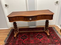 Front and angled view of the antique wooden hall table showing curved kidney-shaped top, single central drawer with glass knob, and turned spindle legs on a red patterned rug.