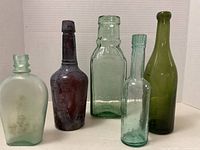 Five antique and vintage bottles in a row against a white background showing front view: clear, dark purple, light green, light aqua, and olive green bottles.