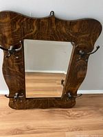 Front view of antique wooden framed mirror with four metal clothes hooks attached to the frame.