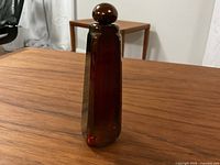 Perfume bottle standing upright on wooden table, showing faceted red glass body and rounded stopper top.