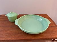 Pie plate and sugar bowl set displayed on wooden surface. Both pieces are pale green glass with the pie plate featuring side handles and the sugar bowl a lid.