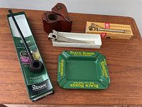 Overview of all four items on wood surface: Peterson Churchwarden pipe in box, wooden tobacco holder with inlay and lid, Solingen cigar cutter in box, Black Horse Ale green enamel ashtray with yellow text.