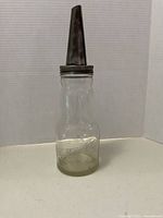 Full view of the vintage glass oil bottle with metal spout standing upright on a flat surface against a plain background.