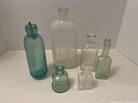 Photo showing six vintage glass bottles including a frosted Underwood Ink bottle, light blue medicine bottle with wire dropper, small green and clear bottles, and a small rectangular light green bottle.