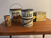 Photo showing five vintage kitchen tins including Jack & Jill peanut butter, Majestic spices, Salada tea, Mother Parker coffee, and Cheese Crispies box on wooden surface.