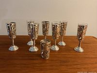Nine vintage sterling silver liqueur and shot glasses arranged on a wooden surface showing hammered silver overlay and varying heights.