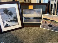 Three ocean-themed pictures, two framed in dark wood, one painting on thick paper, displayed leaning against wall on carpet