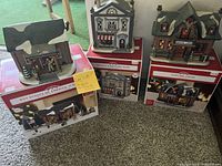 Three ceramic Christmas village buildings on their boxes including Hunting Lodge, Boy Scouts Lodge with figurines, and Delucas Butcher Shop