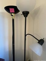 Photo showing two tall floor lamps, one with black torchiere shade and brass detail, the other with frosted white glass shade and adjustable smaller side lamp.