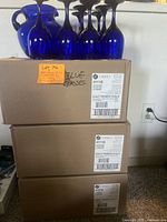 Photo showing blue glass pitcher and several blue wine glasses arranged on cardboard boxes labeled with Libbey brand and model 4111B with contents 12 pieces of Goblet Premiere CB Blue wine glasses.