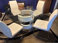 Photo showing 48 inch round glass top dining table with sculptural white and chrome base, surrounded by 4 white faux leather chairs.