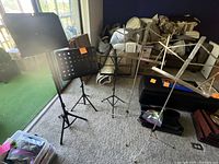Photo showing five music stands of various heights and metal finishes placed on carpet near window and furniture. Visibility of two stands with carrying cases on floor.