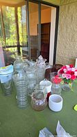 Overview of assorted flower vases and artificial flowers arranged on green tablecloth, including various clear glass and some tinted glass vases, plus small containers.