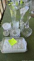 Overview photo showing multiple clear glassware items on green tablecloth including vases, candy dishes, relish plate, and serva-snack tray with note