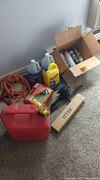 View of assorted automotive items including gas container, jumper cables, tire chains, automotive fluids, and a cardboard box of spray products placed on carpeted floor.