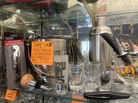 Photo shows shelf display with a hammered stainless steel ice bucket with handle, a polished cocktail shaker, multiple clear shot glasses, a cork-topped wine bottle stopper, bottle pourers, and a packaged black-handled winged corkscrew corkscrew wine opener.