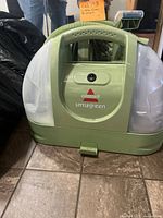 Front view of the Bissell Little Green carpet cleaner showing the power switch, brand, and model name on the green body with clear side tanks.