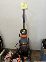 Full view of upright carpet cleaner against a white wall with power cord wrapped around and attachments on floor