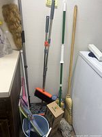 Multiple brooms, mops, and cleaning tools arranged near washing machine and wall.