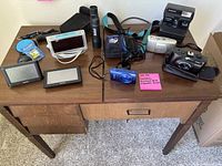 Overview of all items on the wooden desk including cameras, binoculars, Garmin screens, alarm clock, miscellaneous small items with a pink note identifying contents