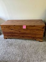 Front view of a wooden cedar chest on carpeted floor showing full chest with natural wood grain and hinged top.