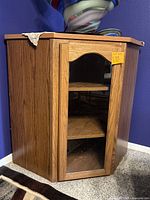 Front view of wooden corner cabinet with glass door showing three shelves inside, positioned in a room corner against purple walls.