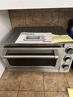 Front angle view of Cuisinart convection toaster oven broiler showing metal body, glass door, handle, and control dials on the right side, placed on a brown countertop against tiled backsplash.