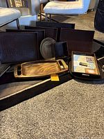 Several trays of different shapes and materials arranged on carpet, visible include a long wooden tray, brown rectangular trays with textured pattern, metallic round tray, black square tray, and wooden picture frame tray.
