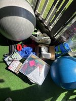 View of entire lot containing the two Pilates balls, plastic storage bin, boxes, and other fitness accessories arranged on artificial grass balcony flooring