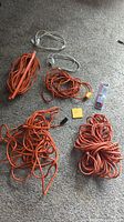 Photo showing five coiled orange extension cords of different lengths laid on carpet with voltage tester in packaging.