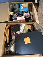 Two cardboard boxes filled with assorted board and card games, including visible titles like Upwords, Patchwork, Doubles 5 Dominoes, and more.