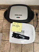 Top view of one George Foreman grill with logo and power indicator lights next to the instruction manual and grill scraper on a tile floor.