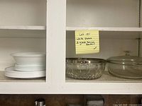 White plates stacked with glass serving bowls on a shelf, including a glass bowl with a silver ring and a glass bowl with lid, showing part of the lot contents.
