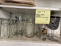 Glass mugs and plastic wine cups arranged on shelf with sticky note description