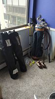 Golf clubs in blue bag alongside hiking poles, Coleman chairs in carrying bags, golf balls, and FootJoy golf shoes on carpeted floor against window and blue wall.